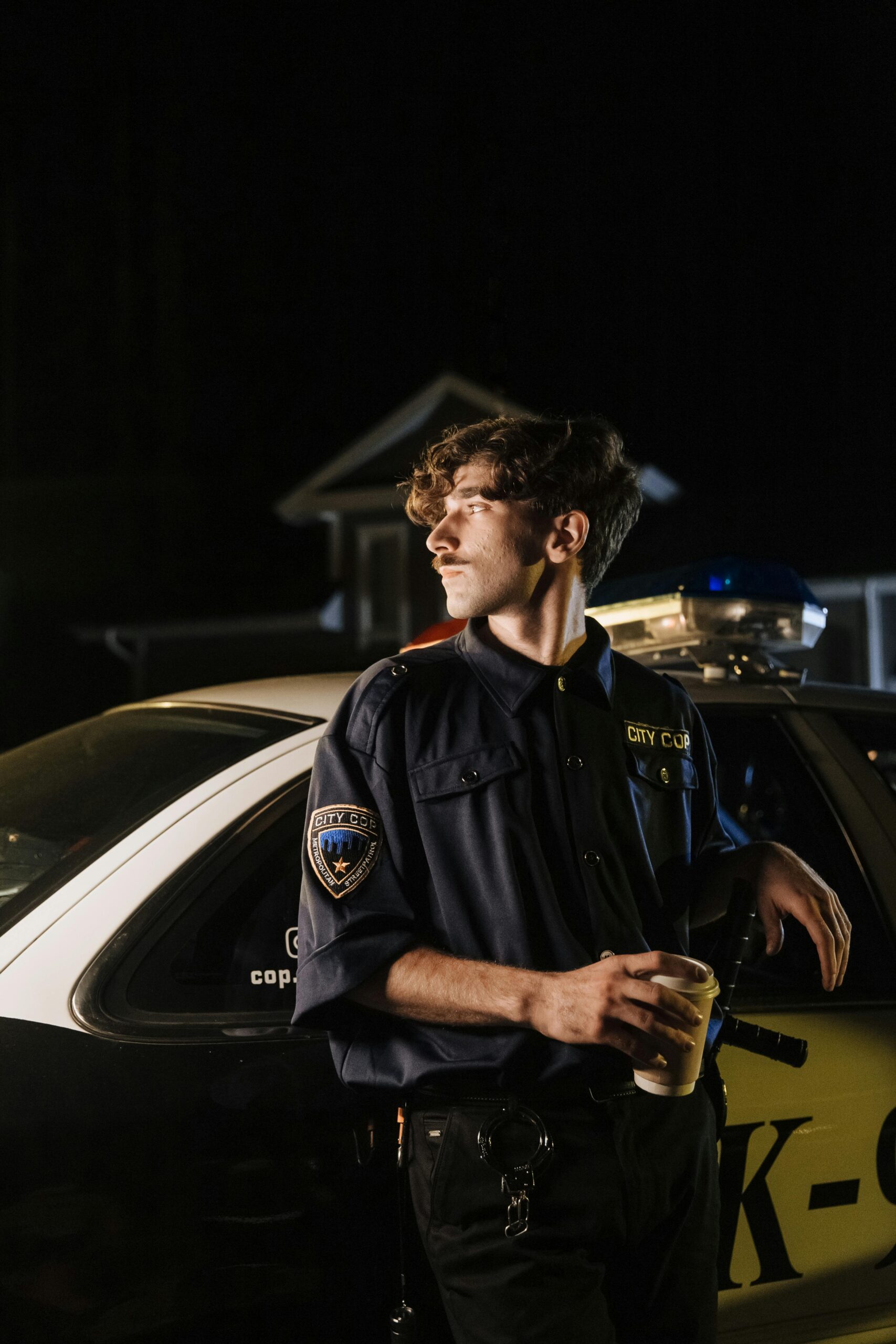 Police officer standing by patrol car at night, holding coffee, looking focused.